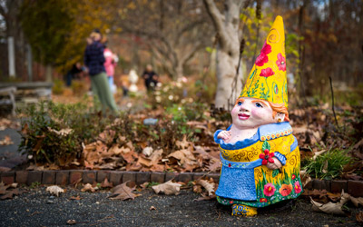 Sylvia the gnome sits in the Nadeau Garden with her yellow floral hat and dress. Behind her, visitors wander among the other gnomes. 